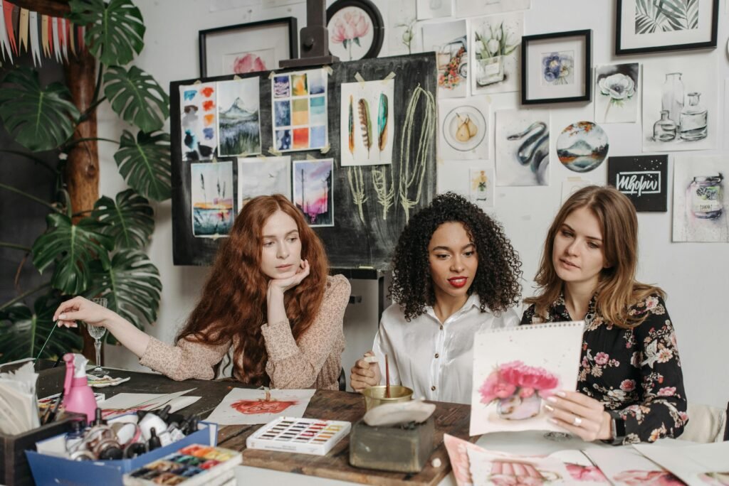 Three women in an art studio discuss watercolor paintings, surrounded by artwork and painting materials.